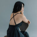 Woman seated on a chair, wearing a black dress with crossed straps on her back, looking to the side.