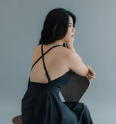 Woman seated on a chair, wearing a black dress with crossed straps on her back, looking to the side.