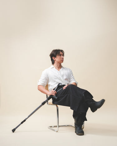 A man sitting on a chair wearing Werable's easy grip buckle shirt with black Hakama culottes pants holding onto a crutch. 