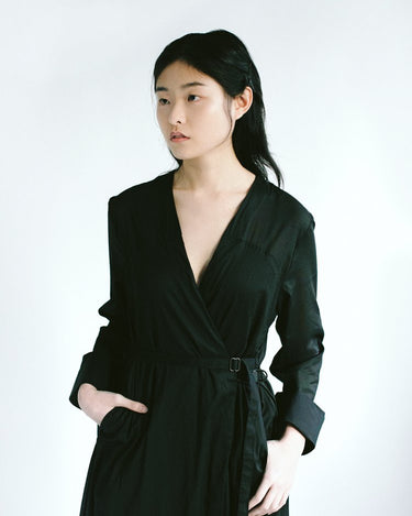 Woman in a black wrap v neck cotton coat with her hands in pockets, looking off to the side.