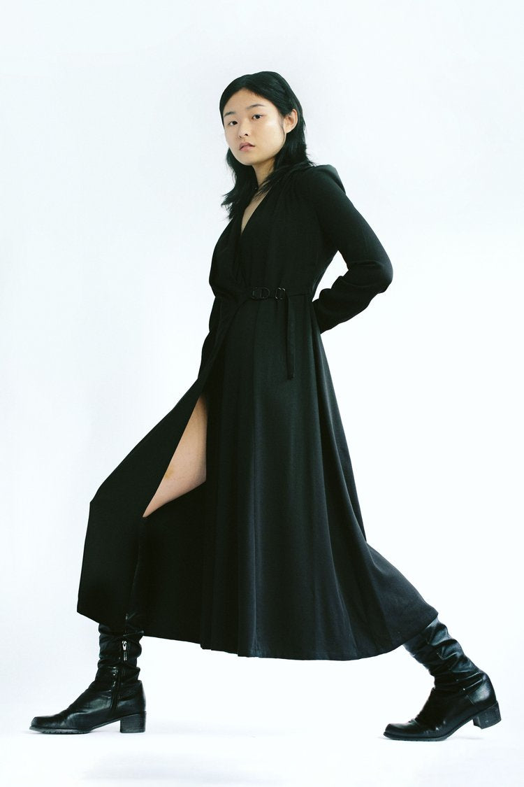 Woman in a black Werable easy grip buckle wrap dress and black boots, posing with one leg extended.