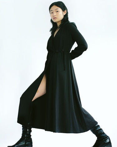 Woman in a black Werable easy grip buckle wrap dress and black boots, posing with one leg extended.