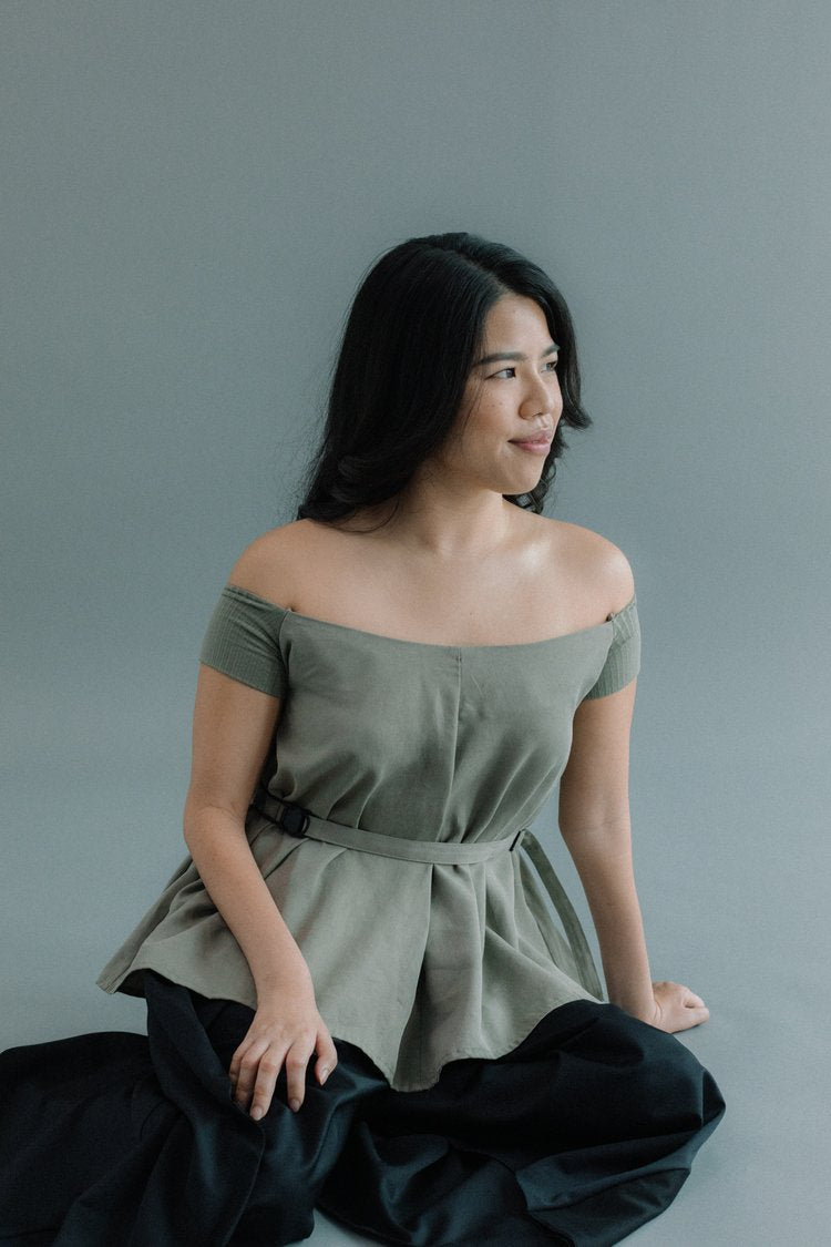 Woman sitting on the floor in a Werable's sage green off shoulder top and hakama culottes in black looking to the side.