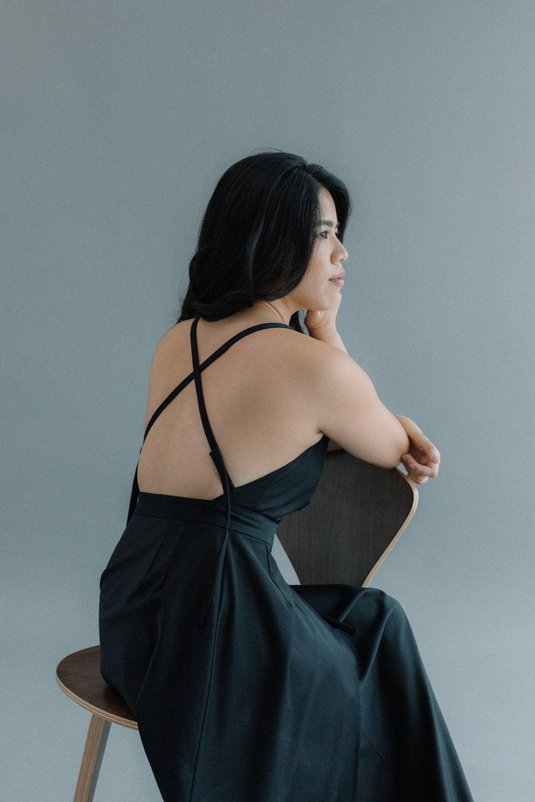 Woman seated on a chair, wearing a black dress with crossed straps on her back, looking to the side.