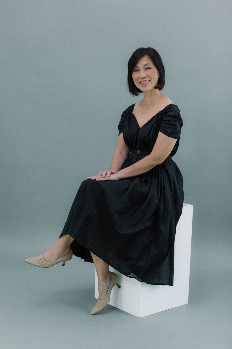 Woman in a black, ruched short-sleeve dress with a V-neckline and a cinched waist, sitting on a white block wearing nude heels.